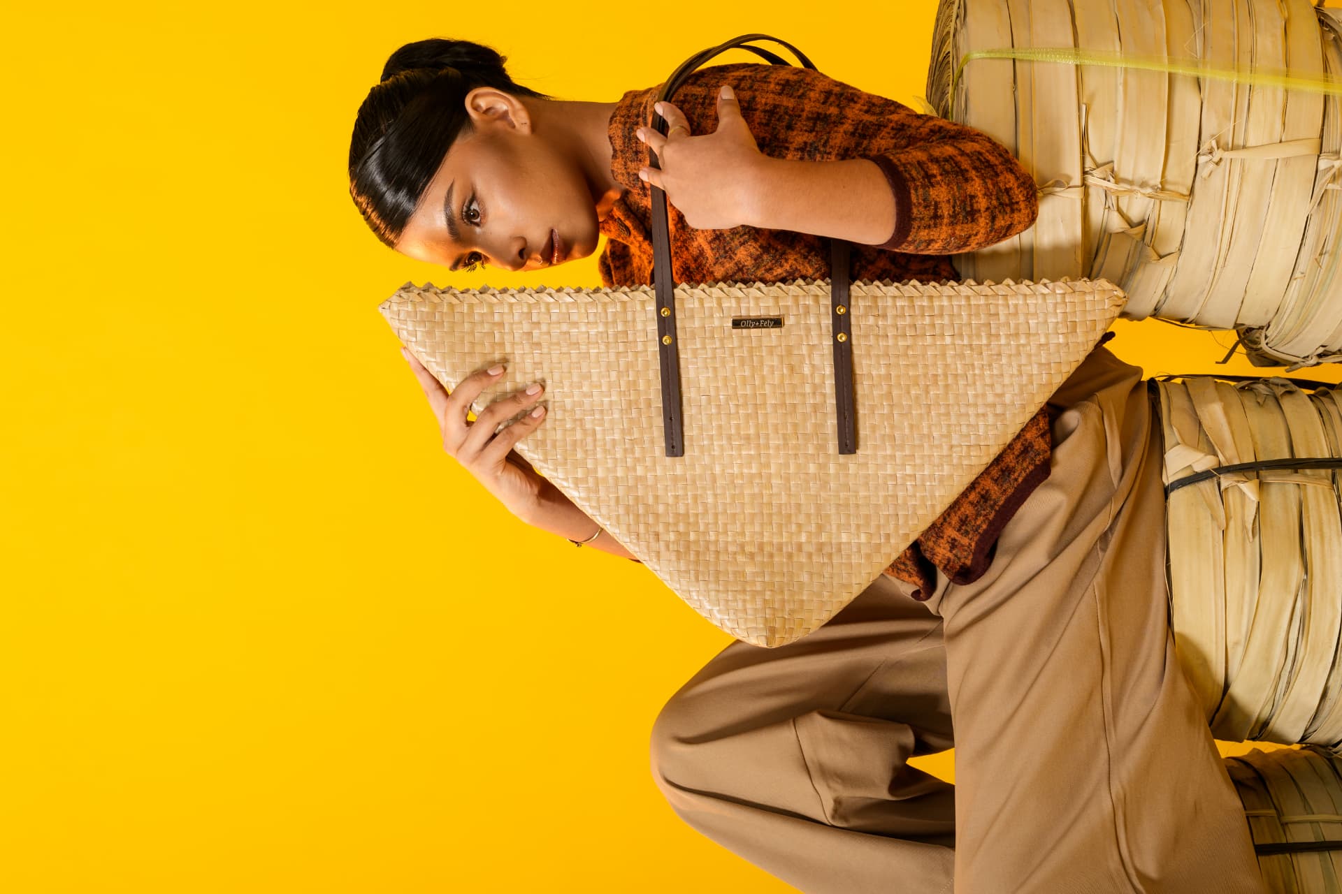 Model in a burnt-orange top holding the natural buri TRIA tote on a yellow backdrop